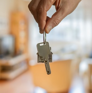 Main tenant un trousseau de clés, incluant une clé et un porte-clés en forme de maison, sur fond flou d'un intérieur résidentiel. Accès au logement.