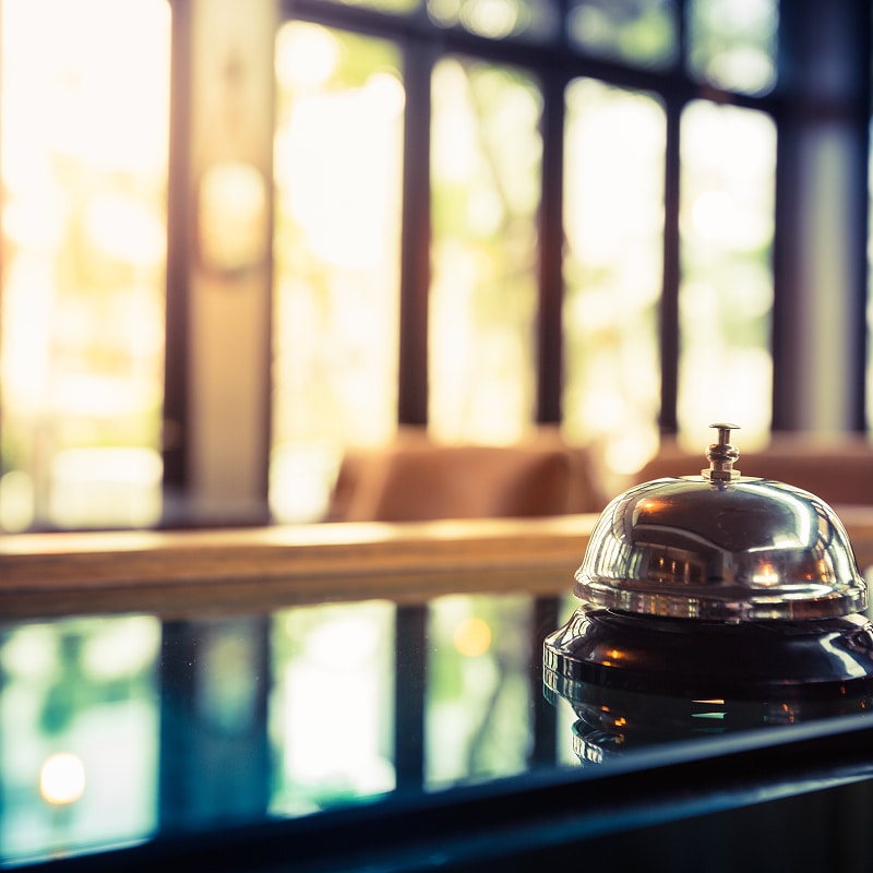 Cloche de service argentée posée sur un comptoir noir brillant, avec un hall d'hôtel lumineux en arrière-plan flou.