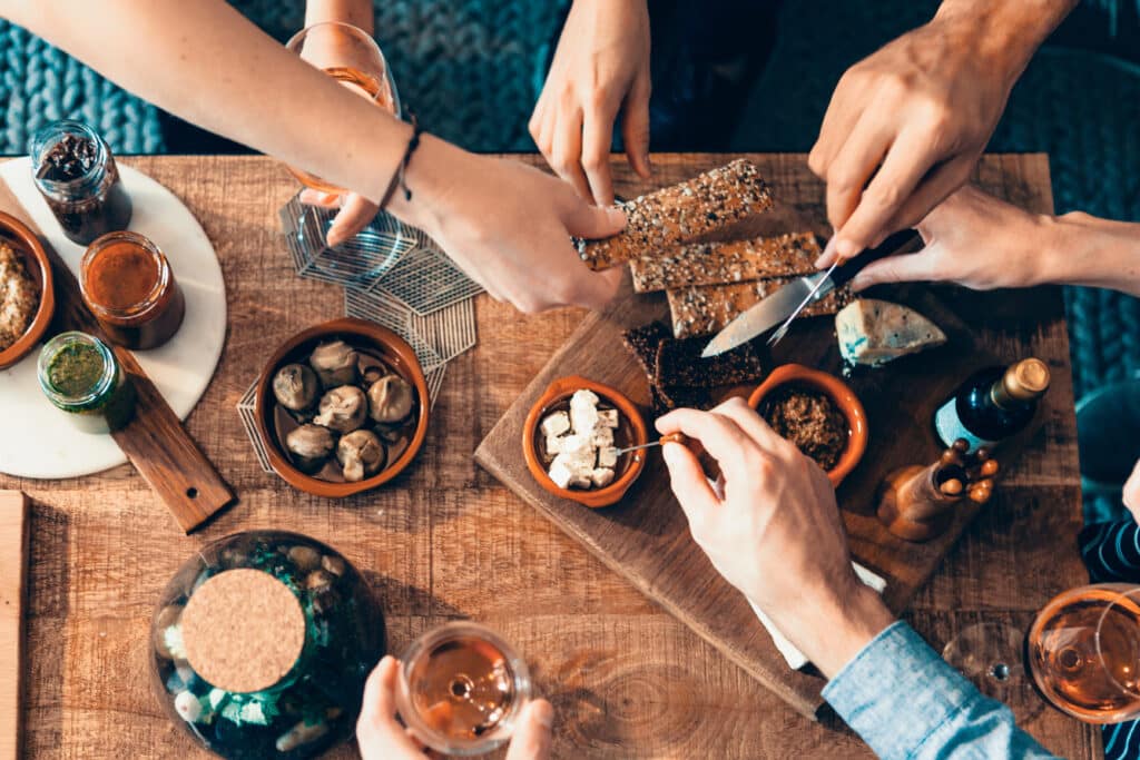 Vue aérienne de mains partageant des craquelins, fromages, sauces, et verres de vin rosé sur une table en bois. Moment convivial.