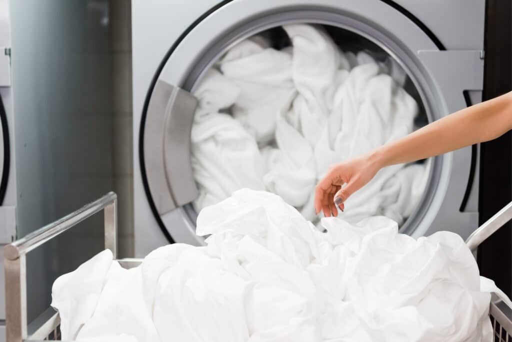 Main saisissant du linge blanc dans un chariot de blanchisserie, avec une machine à laver professionnelle remplie en arrière-plan.
