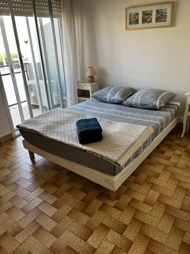 Chambre lumineuse avec un lit double, literie grise à motifs tribaux, et une vue sur un balcon via des portes coulissantes en verre.